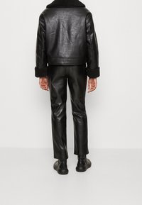 Giacca di pelle nera con colletto in shearling, abbinata a pantaloni in pelle nera aderenti. L'outfit presenta una texture liscia e un design sartoriale.