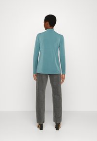 Camisa de manga longa com colarinho em tecido teal suave, combinada com calças cinzentas de cintura alta e corte reto, e sapatos de salto pretos.