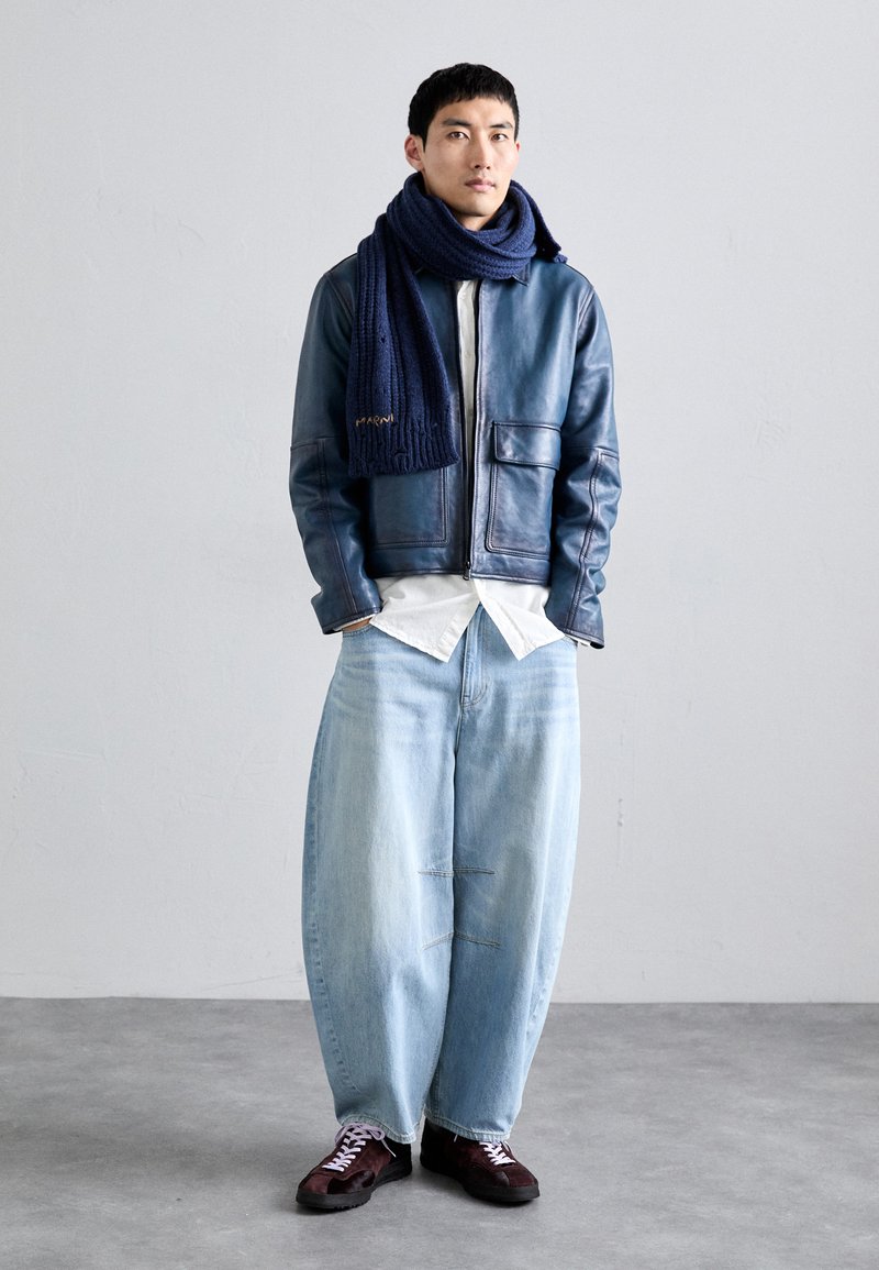 Man staat binnen met een marineblauwe sjaal, blauw leren jasje, wit shirt, brede lichtblauwe jeans en bruine sneakers.