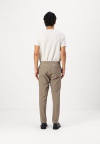 Uomo con capelli ricci scuri in piedi di spalle, indossa una camicia bianca a maniche corte, pantaloni beige a vita elastica e scarpe nere su sfondo bianco.