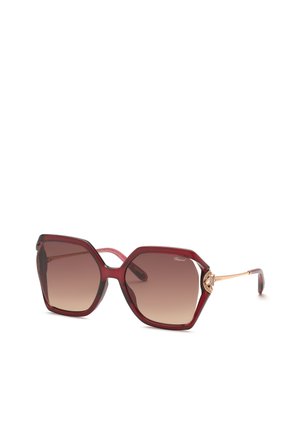 Lunettes de soleil hexagonales surdimensionnées avec montures bordeaux, verres dégradés marron et branches fines dorées avec accents décoratifs près des charnières.