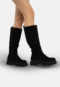 Zwarte dameslaarzen van suède tot over de knie met een stevige zwarte zool, voorzien van een zijrits en een gladde textuur, geschikt voor diverse gelegenheden.