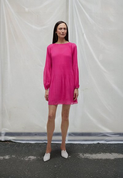 Frau in einem hellpinkfarbenen, langärmeligen Kleid und weißen spitzen Schuhen steht auf Asphalt vor einem weißen halbtransparenten Hintergrund.