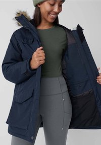 Parka bleumarin cu detalii din blană sintetică, multiple buzunare și captuseală interioară; asortat cu un top verde și colanți gri ajustați.