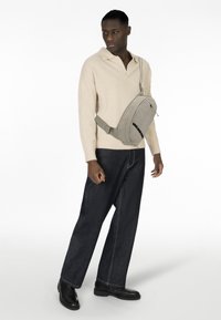 Borsa a tracolla in nylon beige con design curvo, tracolla regolabile e tasca con zip frontale, indossata sopra un maglione crema e pantaloni in denim scuri.