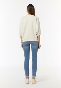 Vrouw met lang bruin haar, gekleed in een crèmekleurige sweater, opgerolde blauwe skinny jeans en beige sneakers, staand met haar rug naar de camera tegen een witte achtergrond.