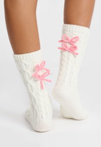 Calcetines de punto trenzado en crema con lazos rosas en la parte de atrás. La tela presenta un patrón texturizado, proporcionando calidez y comodidad.