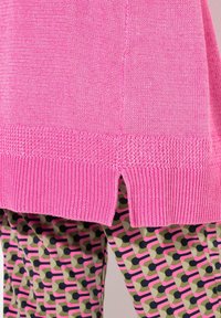 Pull en tricot rose avec un ourlet côtelé et des fentes sur les côtés, associé à des leggings à motifs dans un design géométrique vert, rose et noir.