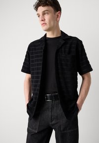 Svart kortermet skjorte med stripet tekstur, åpen krage og frontlomme. Båret over en svart t-skjorte med mørke denimbukser og et svart belte.