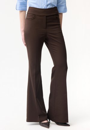 Žena nosící vysoké hnědé rozšířené kalhoty s předními kapsami a černými botami s špičatou špičkou, doplněné světle modrou pruhovanou košilí.