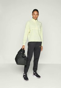 Kvinde i en lys gul zip-up jakke, sorte joggingbukser og sorte sneakers holder en sort træningstaske og står mod en ensfarvet baggrund.