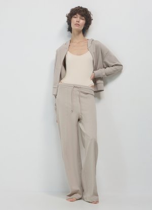 Woman wearing beige tank top, matching loose sweatpants, and a cropped zip-up hoodie, standing barefoot against a white wall.