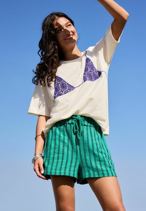 Junge Frau in weißem T-Shirt mit lila Spitzen-BH-Design und grün gestreiften Shorts steht lächelnd vor klarem blauem Himmel.