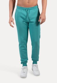 Pantalones de chándal turquesa en material de felpa suave, con cinturilla y puños elásticos, que cuentan con cordón y bolsillos laterales. Llevados con zapatillas blancas.
