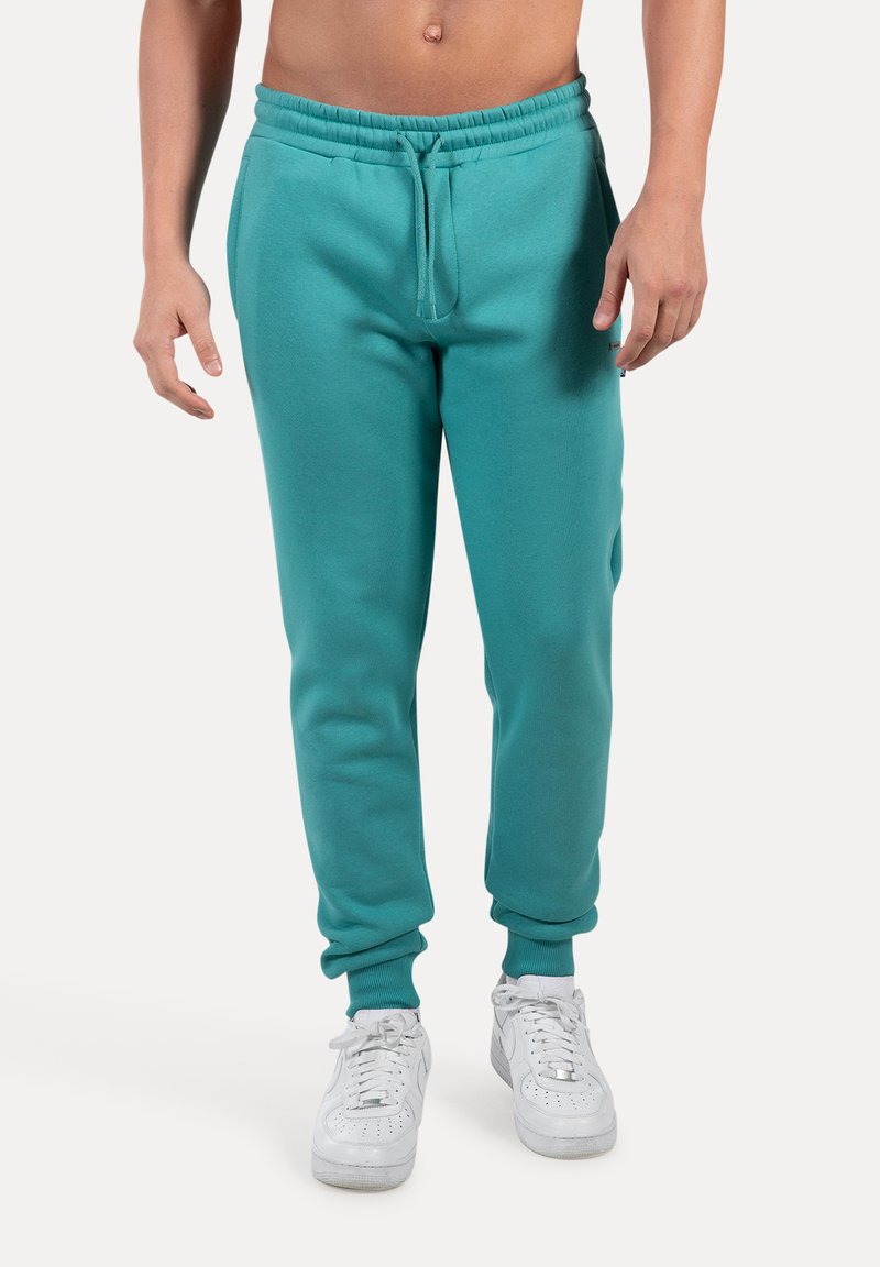 Pantalones de chándal turquesa en material de felpa suave, con cinturilla y puños elásticos, que cuentan con cordón y bolsillos laterales. Llevados con zapatillas blancas.