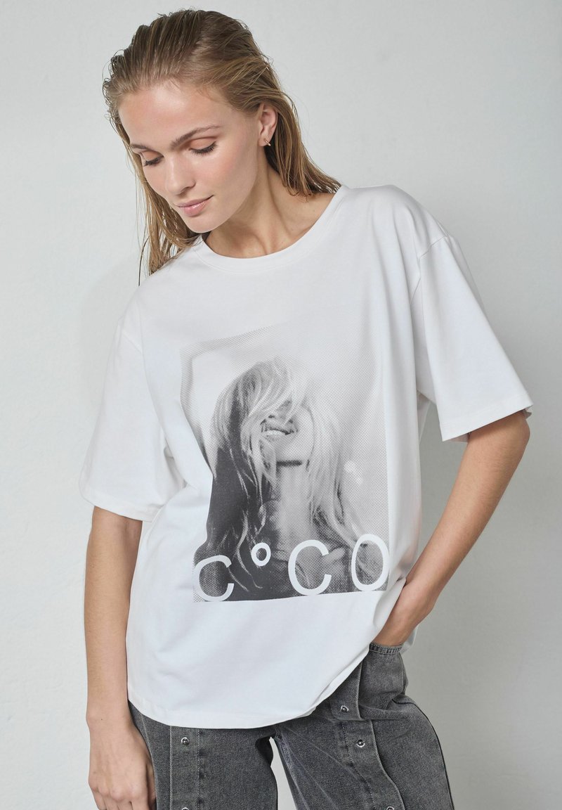 Balta kokvilnas T-krekls ar lielu melnbaltu grafisko attēlu. Īsas piedurknes, brīvs piegriezums ar apaļu kakla izgriezumu un gluda tekstūra.