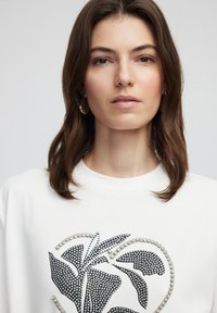 Sudadera blanca con un diseño floral en negro y adornos de rhinestones que delimitan el patrón. Textura suave, cuello redondo.