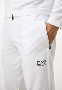 Pantalons de sport blancs avec un motif texturé, des poches zippées et le logo « EA7 Emporio Armani » en noir sur le côté.