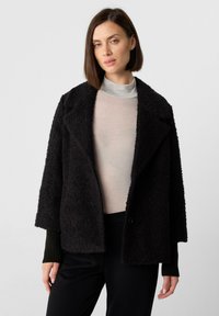 Cappotto nero strutturato con ampi rever e spalle cadenti. Presenta polsini neri a coste e chiusura con un unico bottone. Indossato sopra una maglia chiara.