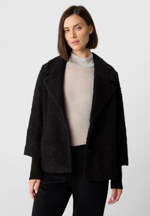 Cappotto nero strutturato con ampi rever e spalle cadenti. Presenta polsini neri a coste e chiusura con un unico bottone. Indossato sopra una maglia chiara.