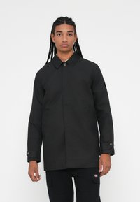 Cappotto esterno nero con colletto, chiusura a bottoni e tasche laterali. Presenta maniche lunghe e un taglio dritto.