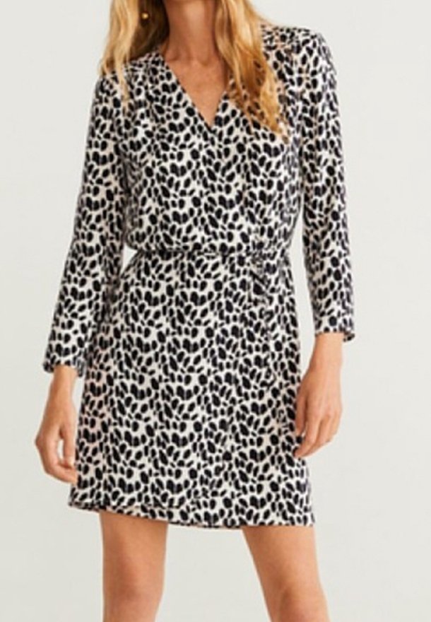 Femme portant une robe portefeuille noire et blanche à pois, manches longues et ceinture à la taille, debout devant un fond uni.