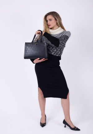 Kvinde i en sort nederdel og grå mønstret sweater, der holder en sort håndtaske, stående foran en hvid baggrund.