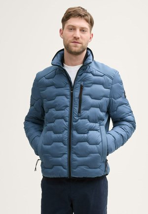 Blauwe pufferjack met een hexagonaal patroon, hoge kraag, ritssluiting aan de voorkant en zijzakken. Stof lijkt glad en lichtgewicht.