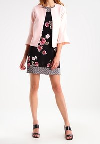 Różowa krótka kurtka na czarnej sukience w kwiaty z wzorkowanym dołem, połączona z czarnymi sandałami na wysokim obcasie. Gładki materiał i rękawy dzwonowe.
