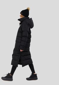 Schwarzer langer Daunenmantel mit Kapuze, ausgestattet mit horizontalen Steppnähten, seitlichen Schlitzen und einer glatten Textur. Getragen mit schwarzen Leggings und Stiefeln.