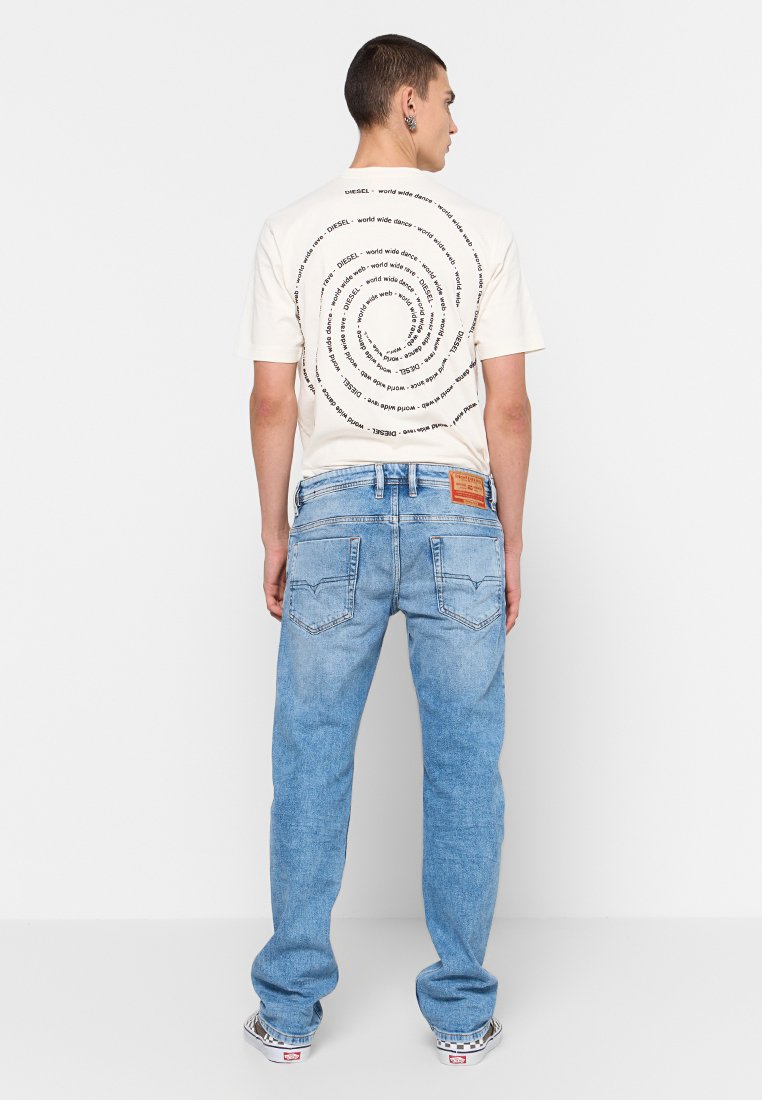 T-shirt crema con design di testo a spirale sulla schiena, abbinata a jeans in denim azzurri. I jeans presentano tasche posteriori e un'etichetta del marchio.