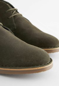 Zapatos de ante verde oscuro con puntera redondeada, costuras mínimas y suela de goma marrón, que destacan por su textura suave y diseño limpio.