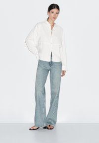 Hvid oversized skjorte med rund krave, kombineret med lyseblå vidde jeans og sorte sandaler, mod en ensfarvet baggrund.