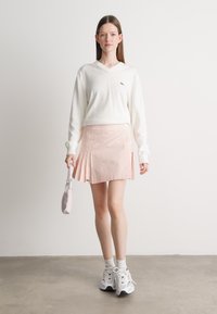 Witte lange mouwen V-hals trui, roze geplooide rok met verticale strepen en witte sneakers. Model houdt een kleine roze tas vast.