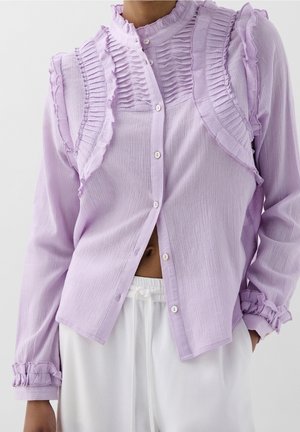 Lila blouse met knopen en verticale ruches aan de voorkant en op de manchetten, gedragen met witte broek met trekkoord.