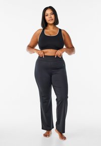 Reggiseno sportivo nero con bordo bianco abbinato a pantaloni neri attillati, entrambi realizzati in un materiale liscio e elastico. La modella è in piedi a piedi nudi, con le mani sui fianchi.