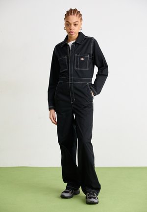 Mono de lona negro con costuras blancas, un collar, dos bolsillos en el pecho y una cremallera frontal. Combinado con zapatillas grises sobre una superficie verde.