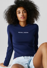 Mlada ženska z kodrastimi lasmi, oblečena v temno modro majico z dolgimi rokavi z napisom "RED BULL RACING" in svetlo modre jeans kratke hlače, sedi pred nevtralnim ozadjem.