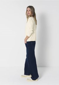 Cremefarbener Strickpullover mit gerippten Bündchen, kombiniert mit dunkelblauen Schlagjeans. Das Model trägt hellfarbige Sneaker und steht vor einem weißen Hintergrund.