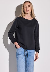 Sudadera negra con cuello redondo, mangas largas y cordón en el dobladillo. Combinada con unos vaqueros de tiro claro para crear contraste.