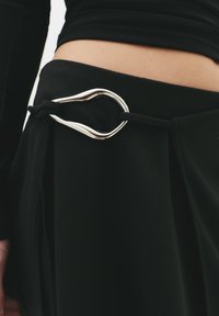 Gonna nera con texture liscia, dotata di una fibbia in metallo argentato a forma di anello allungato, fissata con un nastro di raso nero.