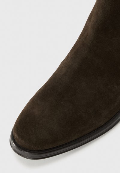 Bottine en suède brun foncé avec un bout pointu, texture douce et semelle en caoutchouc noire. Présente un design épuré et simple avec des accents minimaux.