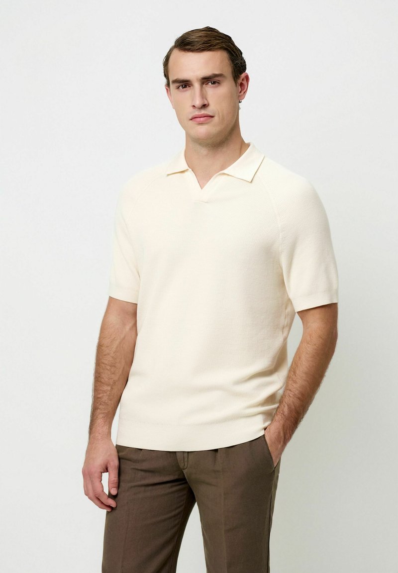 Polo crème à manches courtes avec une texture côtelée et un col en V, associé à un pantalon marron à devant plat. Design simple et moderne.