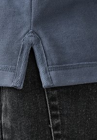 Blå bomullstyg med en strukturerad väv. Har en sidoventilation vid fållen och kontrasterande sömmar längs kanterna. Matcher med svarta denim.