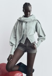 Sivý pletený cardigan s vysokým golierom, prednými gombíkmi a nadrozmernými rukávmi, kombinovaný s hnedými šortkami a čiernymi čižmami po kolená.