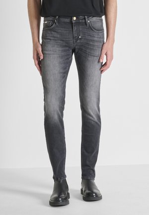 Uomo che indossa jeans neri sbiaditi a taglio slim e stivaletti in pelle nera, in piedi contro uno sfondo chiaro uniforme.