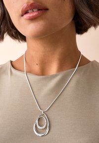 Silver necklace with overlapping teardrop shapes, featuring a textured inner layer and a circular accent, worn on a light brown top.