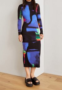 Femme portant une robe ajustée à manches longues, mi-mollet, avec un motif abstrait bleu, vert et rouge, associée à des sandales compensées noires à enfiler.