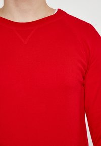 Czerwony sweter z okrągłym dekoltem, o fakturze z prążkowanej dzianiny i rękawach raglanowych. Posiada subtelny detal V-ścisnienia przy szyi.