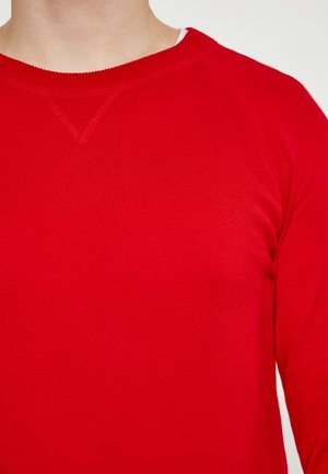 Pulover roșu din lână cu textură ribbed, gât rotund și mâneci raglan. Prezintă un detaliu discret în formă de V la gât.